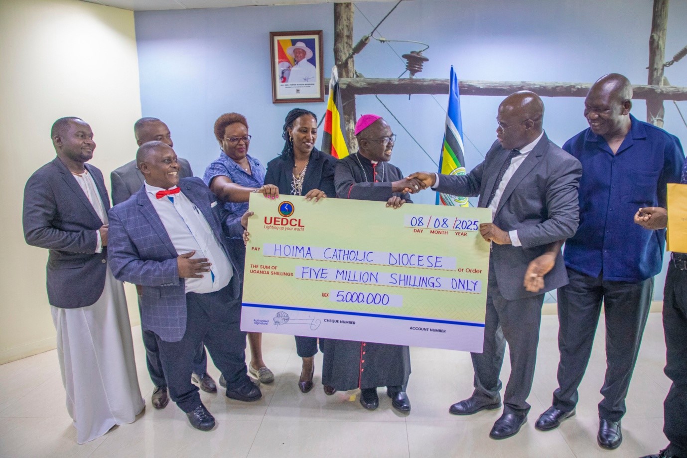 UEDCL Managing Director Paul Mwesigwa (second-right) hands over a dummy cheque for UGX. 5 million to the Bishop of Hoima Catholic Diocese, Rt. Rev. Vincent Kirabo Amooti (sixth-left) and other UEDCL officials on Friday 8th August 2025 at UEDCL Tower.